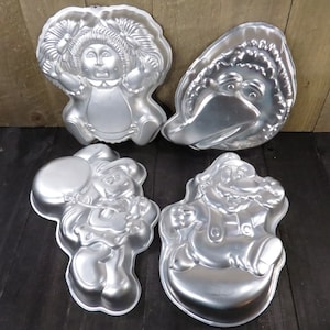 May include: Four silver metal cake pans shaped like cartoon characters. The pans are for baking cakes in the shape of a girl with pigtails, a bird, a cartoon character with a balloon, and a cartoon character with a mustache.