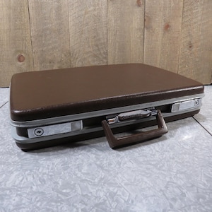May include: A brown leather briefcase with a silver metal handle and clasps. The briefcase has a textured surface and a silver metal nameplate.
