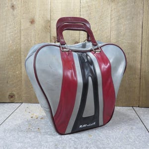 May include: A vintage bowling bag in a gray, red, black, and silver color scheme. The bag has a rounded shape with a burgundy handle and the word "RANGER" printed on the side.