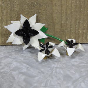May include: A set of three floral-themed jewelry pieces featuring white enameled petals and glossy black centers. The largest piece is a brooch with a green stem and leaf, while the two smaller pieces are earrings with matching white and black designs.