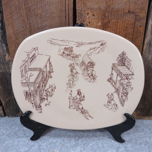 Peut inclure: Un plat ovale en céramique de couleur crème avec un dessin au trait brun représentant une scène de ville de l'Ouest. La scène comprend une diligence, un train de chariots et un saloon avec l'inscription "Fargo Express" sur l'enseigne.