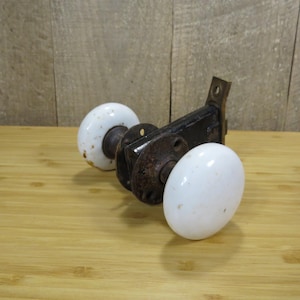 May include: Antique door hardware set featuring a black metal latch and two white porcelain doorknobs. The doorknobs have a classic oval shape and show signs of age, with some discoloration. The set is placed on a wooden surface.