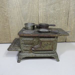 May include: A miniature cast iron toy stove with a weathered green finish and the word EAGLE embossed on the front door. The stove features a flat top with two small metal pots and a pan resting on the burners, supported by four legs.