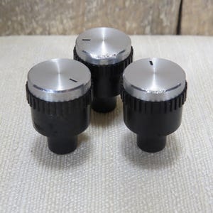 May include: Three silver and black control knobs. Each knob has a silver top with a black line indicator and a black base with a ridged texture. The knobs are designed for electronic devices.