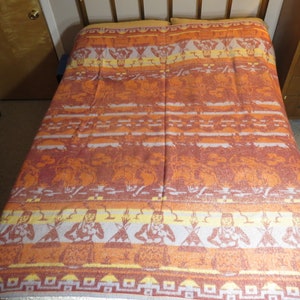 May include: A woven blanket with a Native American design in shades of orange, yellow, and white. The blanket features a repeating pattern of stylised figures and geometric shapes.