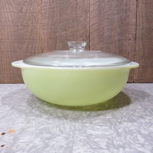 May include: A vintage green glass casserole dish with a clear glass lid. The dish is round and has a white rim.