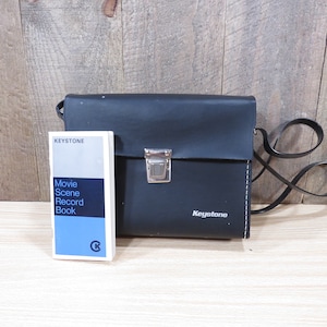 May include: Black leather camera case with a silver clasp and a shoulder strap. The case has the word "Keystone" embossed on the front. A small booklet titled "Keystone Movie Scene Record Book" is next to the case.