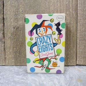 May include: A vintage Crazy Eights card game box with a colorful design. The box features a smiling jester illustration holding a sign that reads "Crazy Eights Card Game." The background is white with colorful polka dots. The box also includes the text "Rule Card Included."