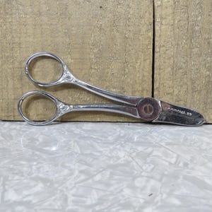 May include: Silver-toned Corona 29 brand scissors with ornate handles. The scissors have a small screw in the center of the handle.