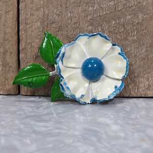 May include: A white flower brooch with blue trim and a blue center. The brooch has green leaves and a silver stem. The flower is set against a wooden background.