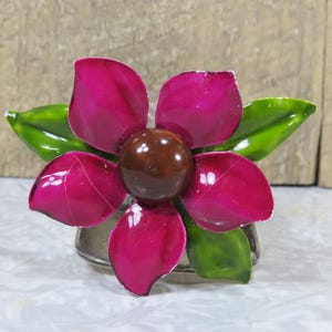 May include: A pink flower brooch with green leaves and a brown center. The flower has five petals and is made of metal.