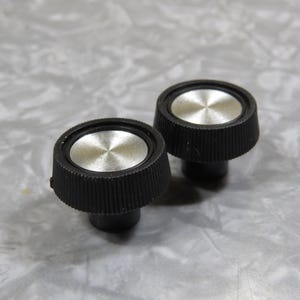 May include: Two black control knobs with silver centers. The knobs have a ridged, conical shape and a circular silver top with a brushed metal finish. The knobs are likely for audio equipment or similar electronics.