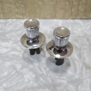 May include: Two chrome knobs, each with a ridged cylindrical top and a flat, circular base. The shiny knobs are designed for a mechanical application. The base is dark and has a unique shape. The knobs are made of metal.
