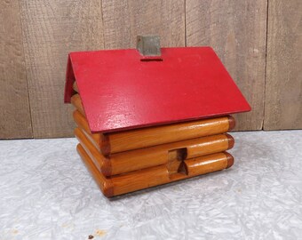 May include: A wooden log cabin piggy bank with a red roof and a slot on top for coins. The cabin is made of stacked logs and has a small chimney on top.