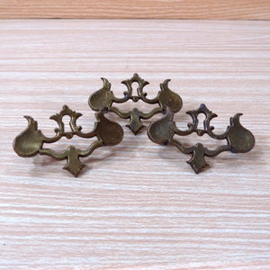 May include: Three antique brass drawer pulls with ornate designs. The pulls have a decorative, intricate, and symmetrical design.