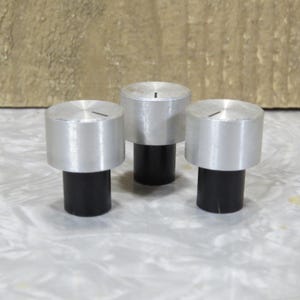May include: Three silver knobs with a black base are shown. Each knob has a cylindrical shape with a flat top and a thin groove. The knobs are evenly spaced and appear to be made of metal. The background is a light brown colour.