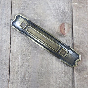 May include: A brass door handle with a geometric design. The handle is rectangular with a raised, textured center and a black border.
