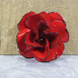 May include: A bright red metal flower brooch with layered, curved petals. Each petal features a distinct, thin black outline along its edge, creating a sharp contrast against the glossy red finish of the flower's surface. The center is dark.