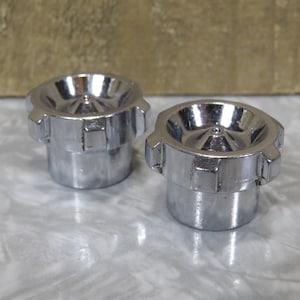 May include: Two shiny, chrome-colored metal objects with a cylindrical base and a flared, bowl-shaped top. The objects have a series of rectangular indentations around the base and are set against a neutral background.