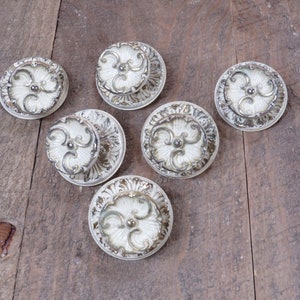 May include: Six white ceramic drawer pulls with ornate silver accents. The pulls have a floral design and are arranged on a wooden surface.