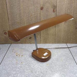 May include: A brown desk lamp with a rectangular shade and a silver-colored stem. The base is oval-shaped with a white on/off switch. The lamp is plugged into an electrical outlet.