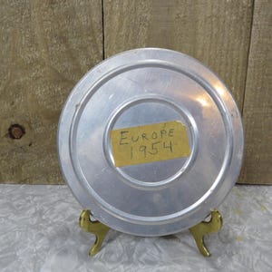 May include: A round, silver-colored metal container with a label that reads "Europe 1954." The container is resting on a small, gold-colored stand. The background is a wooden surface.