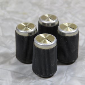 May include: Four black ribbed control knobs with gold and silver detailing. The knobs are cylindrical with a slotted top. These are likely replacement parts for audio equipment or other electronics.