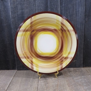 Peut inclure: Un grand plat de service rond en céramique avec un bord brun et un motif tourbillonnant de jaune et de brun. Le plat est sur un support en métal doré.