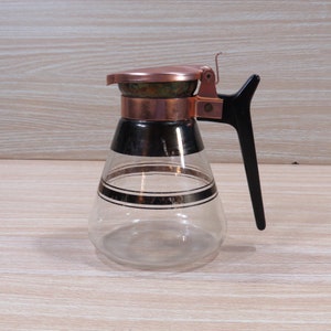 May include: A glass coffee pot with a copper lid and a black plastic handle. The pot has a black and gold stripe design.