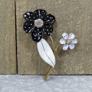 May include: A floral brooch featuring a large black flower with white polka dots and a clear rhinestone center. A smaller white flower with black dots and a rhinestone center is also present. The brooch has a white leaf and gold-tone stem.