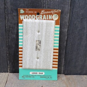 May include: A vintage package for a light gray woodgrain single-pole light switch plate. The package is labeled "The Richness of Beautiful Woodgrain" and "Limed Oak". The package also includes the text "Available in Limed Oak, Walnut, Maple or Birch" and "Bell Electric Co. Chicago 36, Ill."