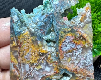 May include: A close-up of a polished, rectangular gemstone with a complex pattern of colors. The stone features shades of blue, teal, yellow, orange, and white, with a rough, textured surface and natural formations. The gemstone is a unique, natural specimen.