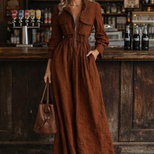 May include: A long, rust-colored dress with a zip-up front, pockets, and long sleeves. The dress is paired with a matching handbag and sandals. The background features a bar setting with vintage signage.