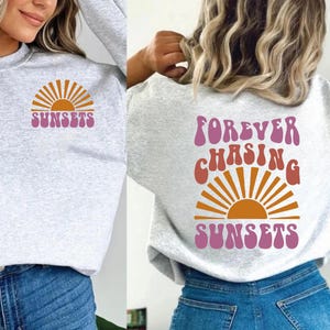 May include: A grey sweatshirt with a pink and orange sunset graphic and the text "SUNSETS" on the front. The back of the sweatshirt has the text "FOREVER CHASING SUNSETS" in a pink and orange retro font with a sunset graphic.