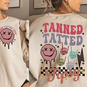 May include: A beige sweatshirt with a graphic design on the front and back. The front design features a pink smiley face with dripping paint and the text "Tanned Tatted Tipsy". The back design features a pink smiley face with dripping paint, a checkered pattern, stars, and the text "Tanned Tatted Tipsy".