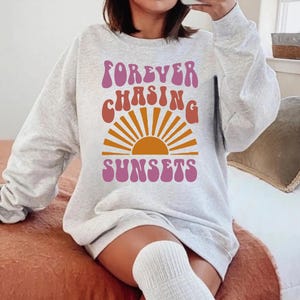 May include: A grey sweatshirt with the text "FOREVER CHASING SUNSETS" in a retro font. The text is in pink, orange and yellow. A stylised sun graphic is behind the text.