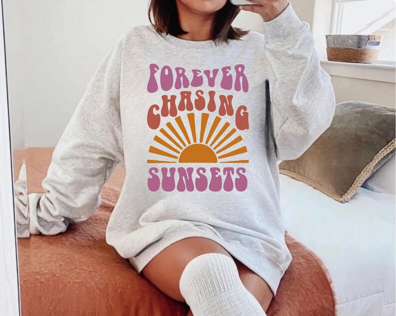 May include: A grey sweatshirt with the text "FOREVER CHASING SUNSETS" in a retro font. The text is in pink, orange and yellow. A stylised sun graphic is behind the text.