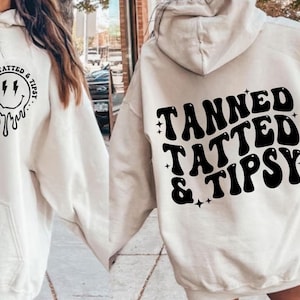 Puede incluir: Dos sudaderas con capucha blancas con texto negro. Una sudadera tiene una cara sonriente con rayos y el texto "Tanned, Tatted & Tipsy". La otra sudadera tiene el texto "Tanned, Tatted & Tipsy" en una fuente retro.