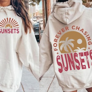 Op de afbeelding: Twee witte hoodies met een grafische print van een zonsondergang en palmbomen. De ene hoodie zegt "Sunsets" en de andere zegt "Forever Chasing Sunsets".