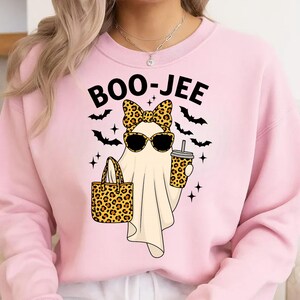 May include: Pink sweatshirt with the text "BOO-JEE" above a cartoon ghost wearing sunglasses and a leopard print bow, holding a leopard print tote bag and a coffee cup. Black bats and stars are also featured.