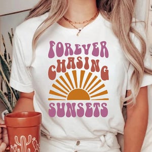 May include: White t-shirt with a pink and orange graphic that says "FOREVER CHASING SUNSETS". The graphic features a stylised sun with rays.