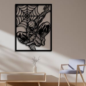 May include: Black metal wall art featuring a silhouette of Spiderman swinging from a web. The art is framed in a black square frame.