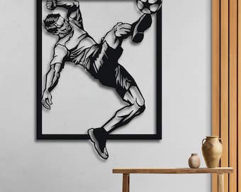 May include: Black metal wall art sculpture of a soccer player kicking a ball. The player is in mid-air, with their leg extended and their foot about to make contact with the ball. The sculpture is framed in a black square.
