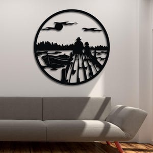 Op de afbeelding: Zwarte metalen wandkunst met een silhouet van twee figuren die over een houten pier naar een boot op een meer lopen. De scène is ingelijst in een cirkel met een maan en wolken op de achtergrond.