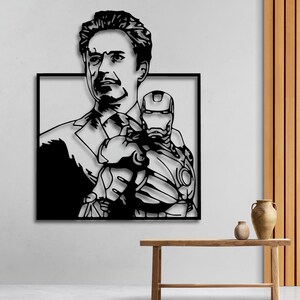 Può includere: Arte da parete in metallo bianco e nero con la silhouette di un uomo che tiene un supereroe tra le braccia. L'uomo ha un'espressione seria e il supereroe indossa un casco.