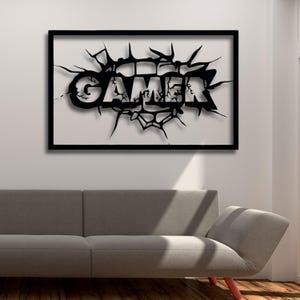Peut inclure: Décoration murale en métal noir avec le mot "GAMER" dans un style fissuré et brisé. L'œuvre est encadrée dans un cadre carré noir.