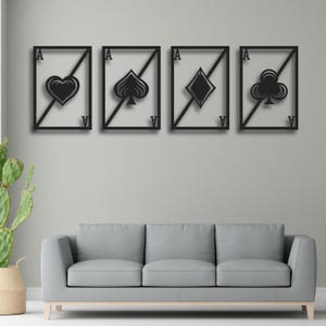 May include: Four black metal wall art pieces depicting the aces of playing card suits: hearts, spades, diamonds, and clubs. Each card is framed with a diagonal line and the letter "A" in each corner. The artwork is displayed above a gray sofa.