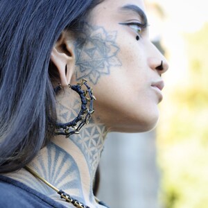 May include: A side profile view of a person with dark hair and intricate face and neck tattoos. The person is wearing large, dark blue hoop earrings with gold-colored accents and a matching necklace with a geometric pendant.