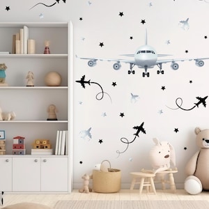 May include: A white wall decorated with airplane and star wall decals. A large airplane decal is centered, with smaller airplane and star decals scattered around. A white bookshelf and children's toys are in the room.