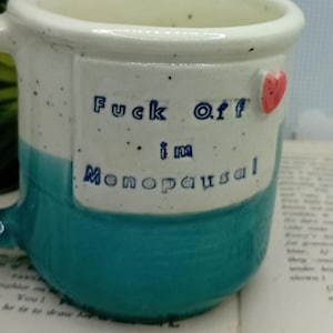 May include: A two-toned ceramic mug with a speckled cream upper section and a solid teal lower section. The cream area features the blue text "Fuck Off i'm Menopausal" and a small, raised red heart accent on the right side of the lettering.